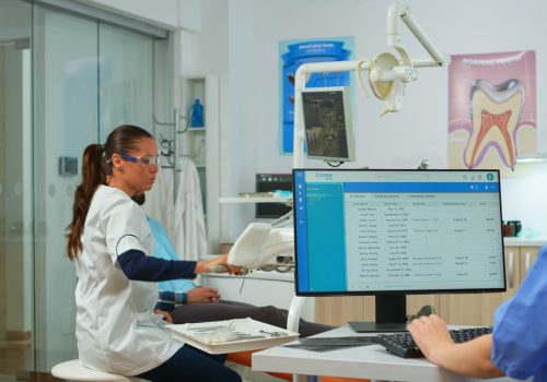 Nurse typing on computer, making appointments while dental specialist talking patient in stomatology chair before examination. Dentist and nurse working together in modern stomatological clinic